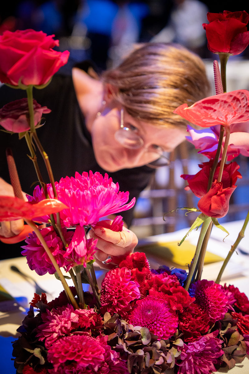 Nahaufnahme einer Tischdekoration mit roten Blumen auf der Bühne der Bayerischen Staatsoper. Im Hintergrund eine Bahlmann Mitarbeiterin, die die letzten Handgriffe am Blumenschmuck macht.
