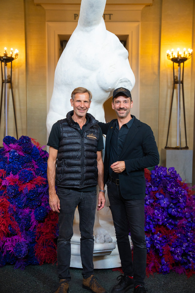 Die Bahlmann-Geschäftsführer Steffen Laible und Michael Kötzel vor einer Marmorstatue in der Bayerischen Staatsoper. Um die Statue sind Bahlmann Blumenarrangements in satten Rot-, Pink- und Lilatönen platziert.