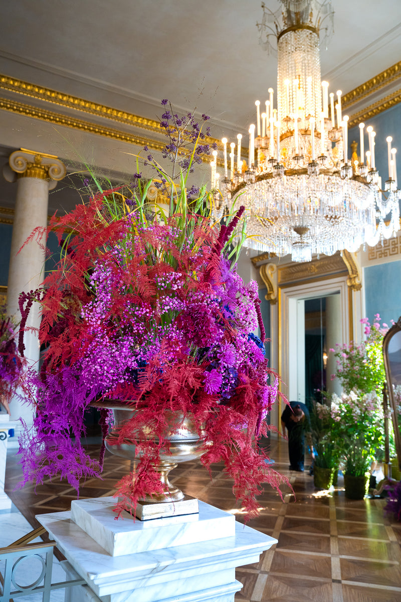 Opulentes Blumenarrangement im Foyer der Bayerischen Staatsoper, mit prunkvollem Kronleuchter im Hintergrund.
