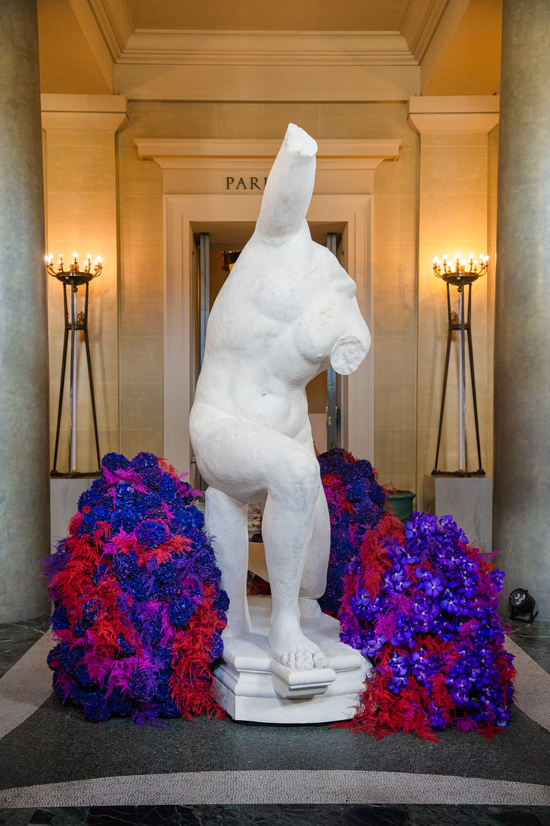 Opulente Bahlmann Blumendekoration im Eingangsbereich der Bayerischen Staatsoper zum Benefiz-Bühnendinner, mit Marmorskulptur in der Mitte, umringt von roten und blauen Blüten von Bahlmann.