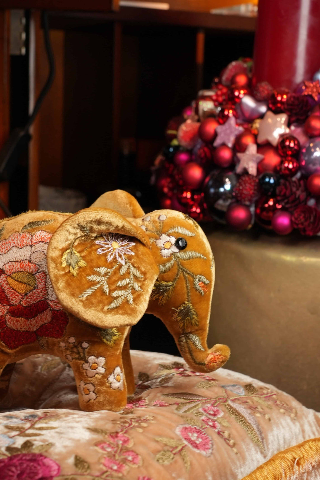 Ein Bahlmann Kuscheltierelefant in Gold, der auf einem Bahlmann Kissen platziert wurde. Im Hintergrund ist ein roter Adventskranz zu sehen.