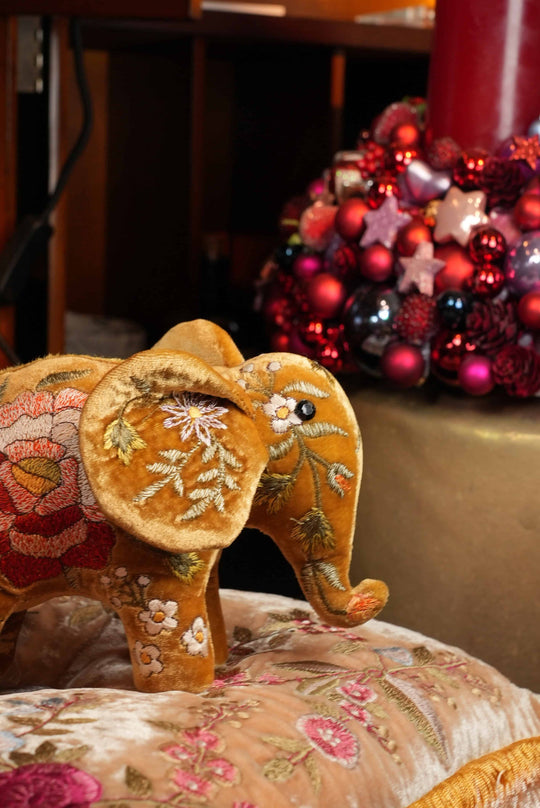 Ein Bahlmann Kuscheltierelefant in Gold, der auf einem Bahlmann Kissen platziert wurde. Im Hintergrund ist ein roter Adventskranz zu sehen.