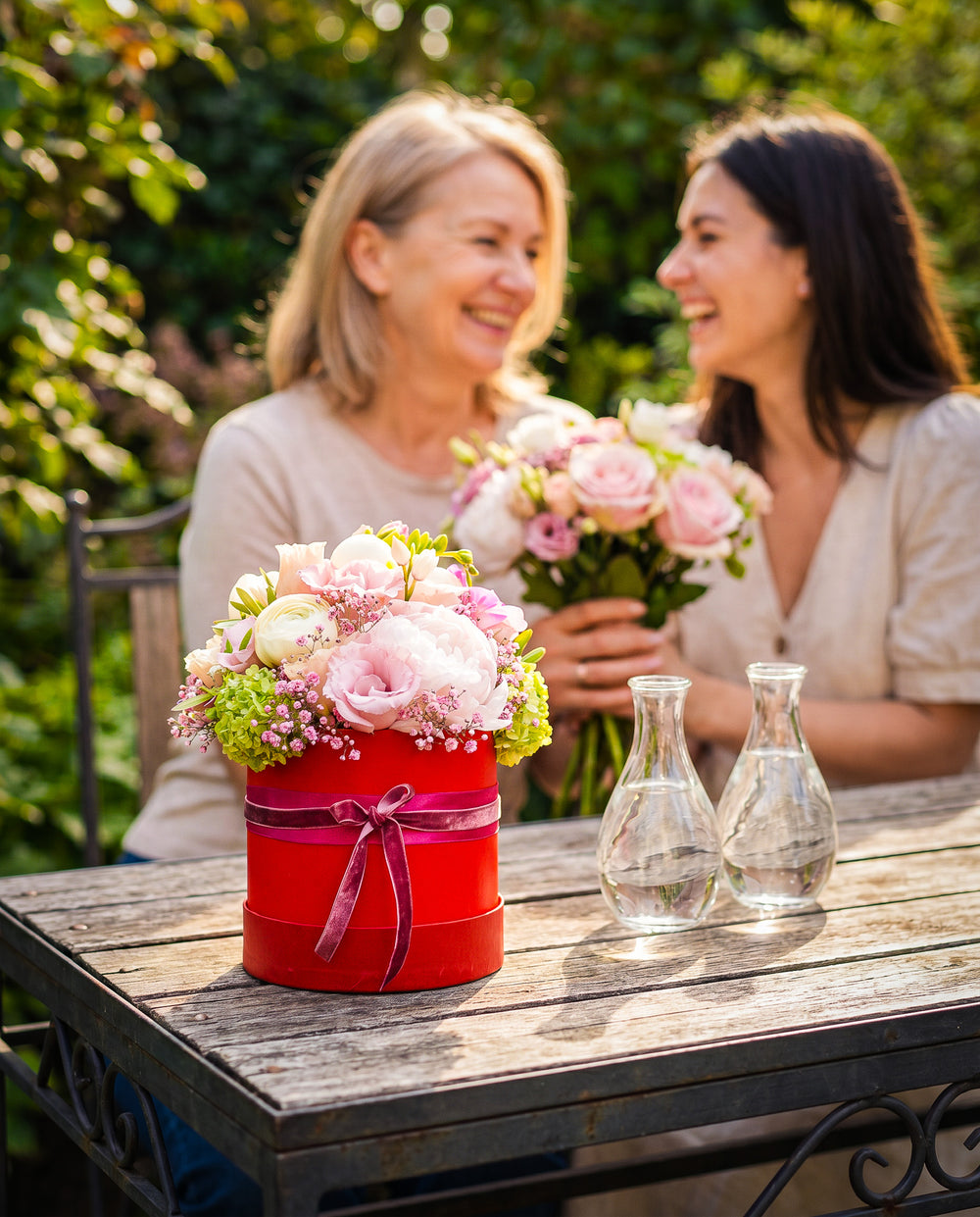 Blumengesteck Love Box in Rot von Blumen Bahlmann in München. Eine rote Geschenkbox, die nach oben hin offen ist und mit unterschiedlichen Blumen wie Rosen, Pfingstrosen und Hortensien gesteckt ist. Das ganze Jahr erhältlich aber besonders beliebt zu Valentinstag und Muttertag. Im Hintergrund sehen wir Mutter und Tochter zum Muttertag, wie die Mama ein Blumenstrauß von der glücklichen Tochter erhält.