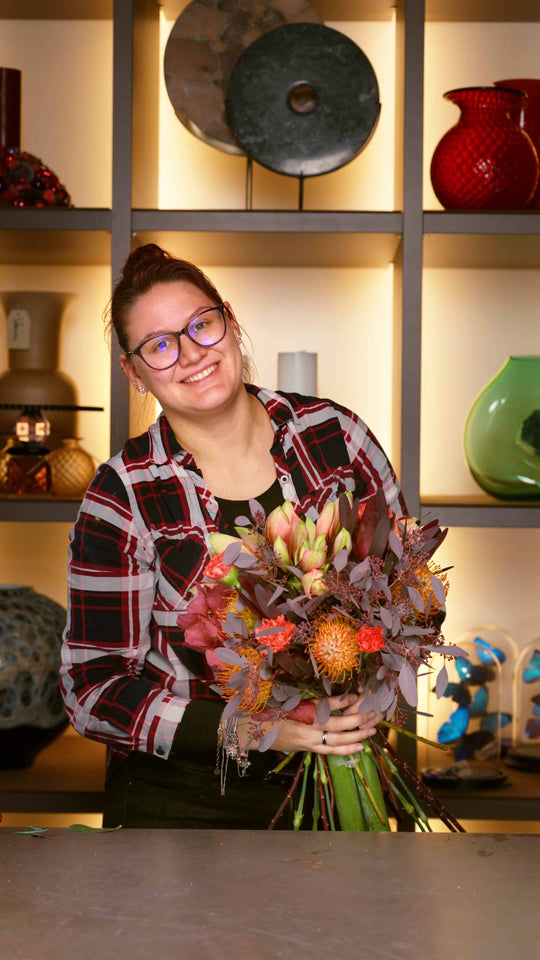 Bahlmann Mitarbeiterin im SHOWROOM in München hält einen Winterstrauß in in die Kamera, bestehend aus Amaryllis, Nadelkissen, Eukalyptus, Orchideen, Nelken und Kapgrün.