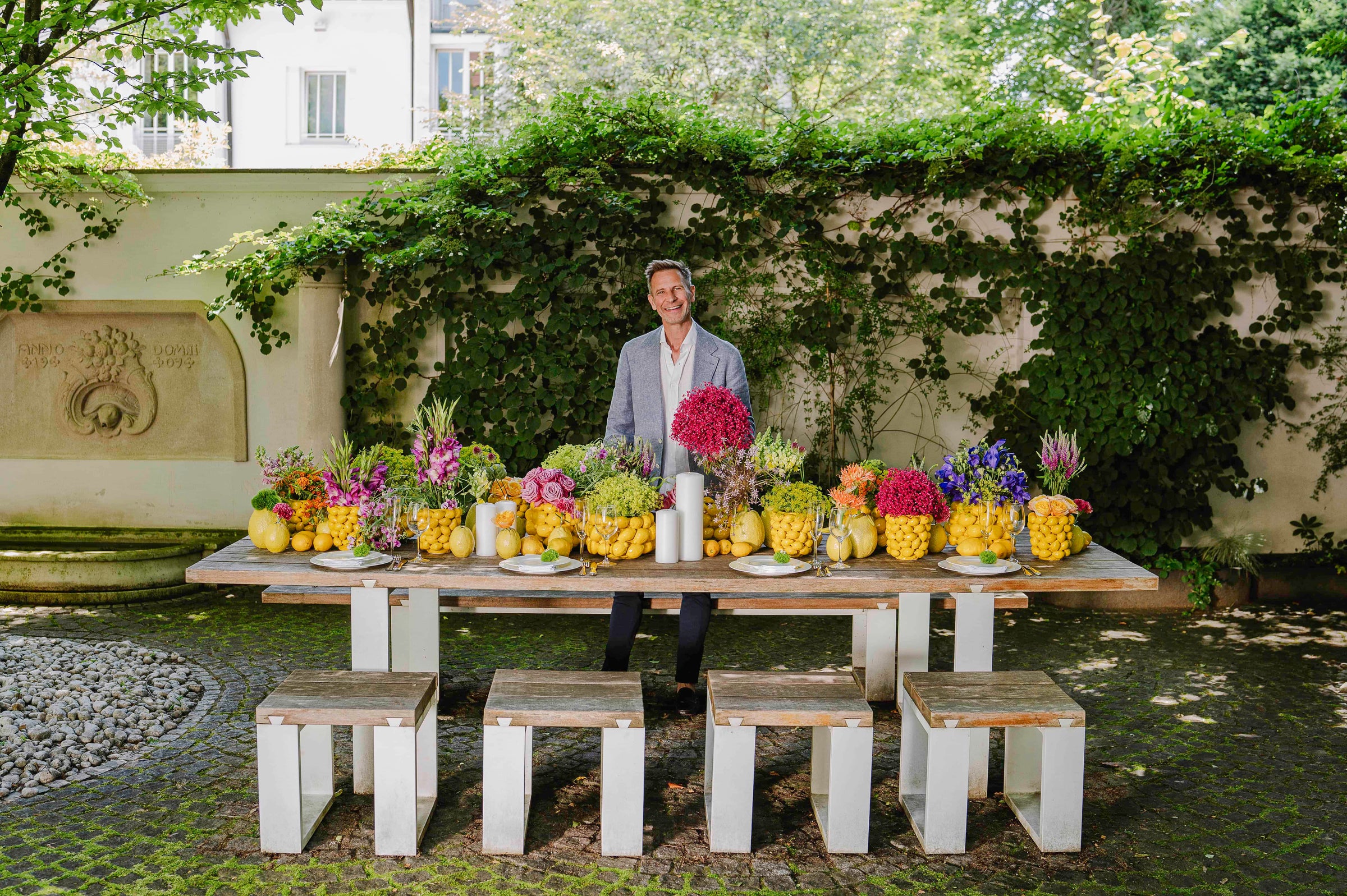 Blumen Bahlmann Geschäftsführer Michael Kötzel steht vor einem sommerlich arrangierten Tisch im Garten mit Zitronen- und Limettenvasen von Bahlmann München.