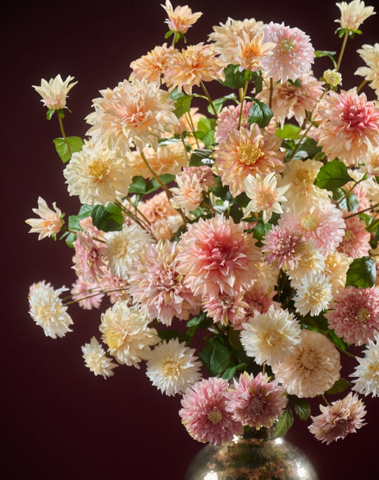 Elegantes Kunstblumengesteck aus pastellfarbenen Dahlien mit grünem Blattwerk vor dunkelrotem Hintergrund. Blumen Bahlmann München.