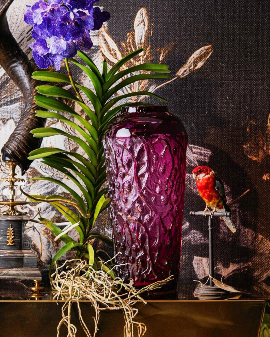 Eine große pinkfarbene Vase von Lalique, präsentiert auf einem gold-verspiegelten Tisch von Bahlmann. Zusammen auf dem Display ein ausgestopfter Wellensittich und aufwendig verzierte Gnu-Hörner auf Marmorsockeln.