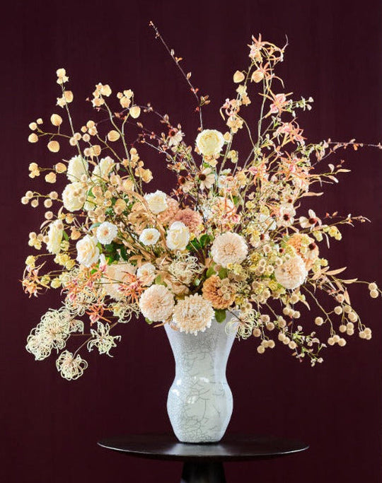 Üppiges Seidenblumenarrangement in Creme, Champagner, Apricot und Rosé mit filigranen Zweigen in einer hellgrauen Vase. Blumen Bahlmann München.