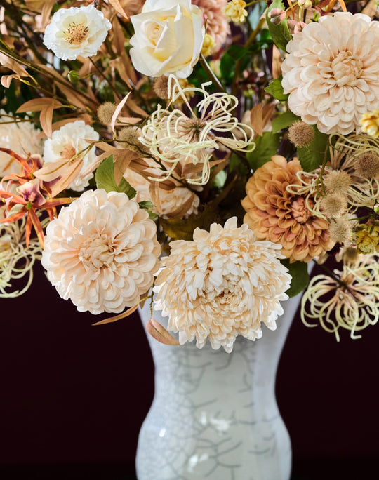 Elegantes Kunstblumengesteck mit cremefarbenen Blüten, feinen Ranken und locker aufragenden Zweigen in einer hohen Vase. Blumen Bahlmann München.