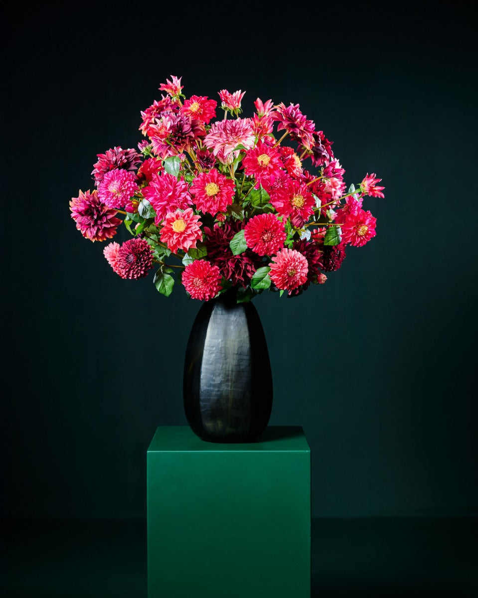 Üppiges Seidenblumenarrangement aus roten, pinken und bordeauxfarbenen Dahlien in einer hohen schwarzen Vase. Blumen Bahlmann München.