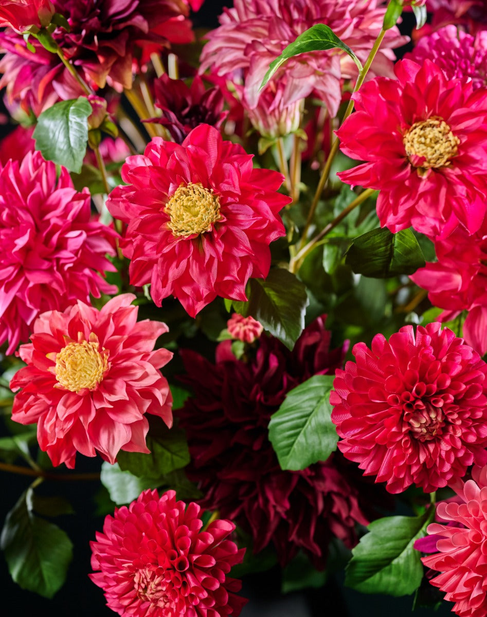 Großes künstliches Dahlienarrangement in kräftigem Rot und Fuchsia mit grünem Blattwerk vor dunklem Hintergrund. Blumen Bahlmann München.