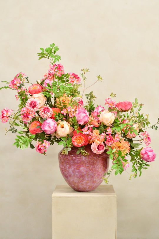 Opulentes Seidenblumenarrangement in Pink, Rosa, Koralle, Apricot und Creme mit lockerem Grün in einer schimmernden rosé-violetten Vase. Blumen Bahlmann München