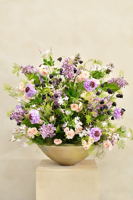 Üppiges Seidenblumenarrangement in Flieder, Violett, Rosé und Creme mit dunklen Akzenten in einer beigefarbenen Schale. Blumen Bahlmann München