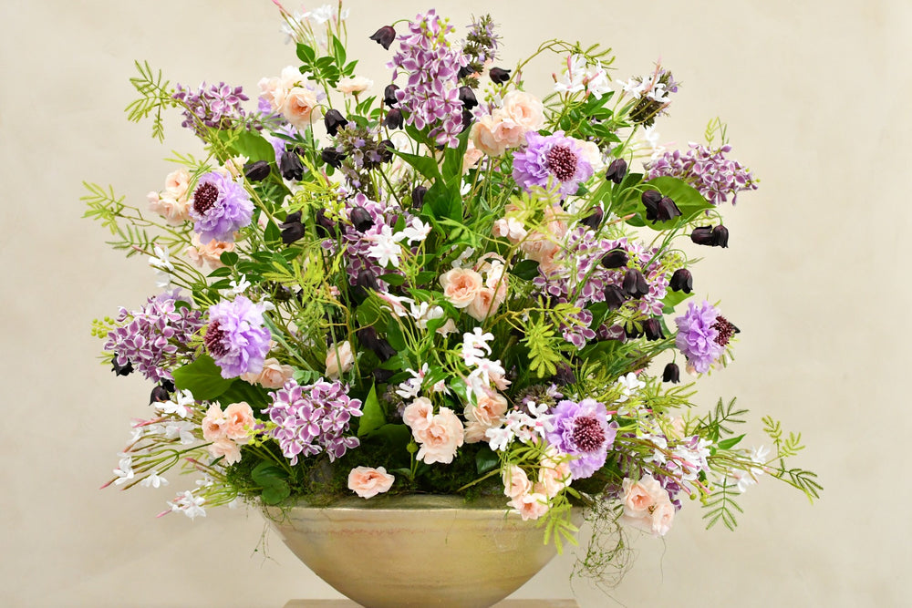 Großes künstliches Blumenarrangement mit violetten Blüten, zarten Roséblüten und locker auslaufendem Grün in einer breiten Vase. Blumen Bahlmann München