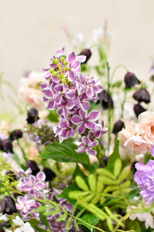 Elegantes Seidenblumengesteck mit fliederfarbenen, cremefarbenen und dunklen Blüten sowie filigranen grünen Zweigen. Blumen Bahlmann München