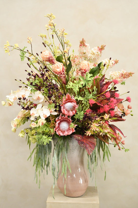 Großes Seidenblumenarrangement in Rosé, Koralle, Bordeaux und Creme mit exotischen Blüten und herabhängendem Grün in einer hohen Vase. Blumen Bahlmann München