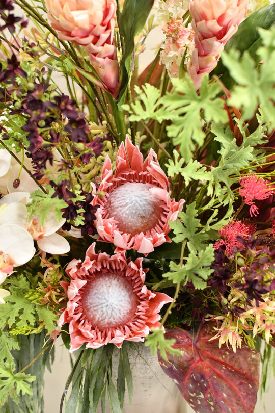 Opulentes Kunstblumengesteck mit Protea, weißen Orchideen, rosa Blüten und dunkelroten Blättern in einer roséfarbenen Vase. Blumen Bahlmann München.
