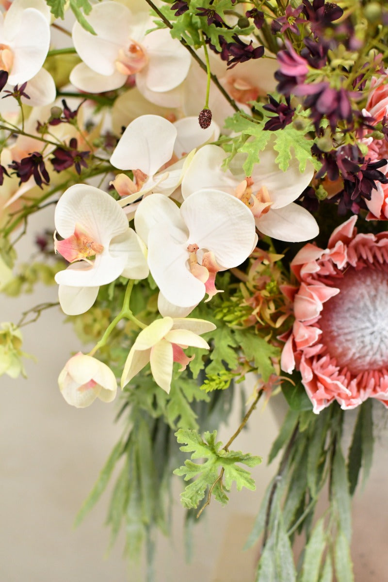 Exotisches Seidenblumenarrangement mit markanten Blüten, filigranen Zweigen und üppigem Grün in einer hohen glänzenden Vase. Blumen Bahlmann München.