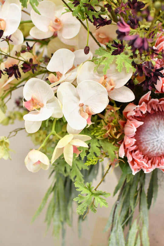 Exotisches Seidenblumenarrangement mit markanten Blüten, filigranen Zweigen und üppigem Grün in einer hohen glänzenden Vase. Blumen Bahlmann München.