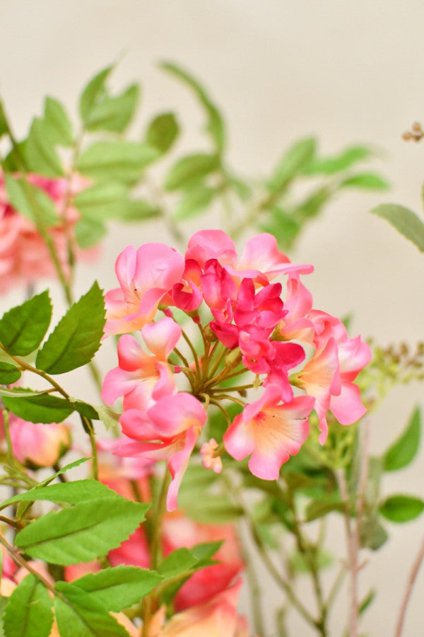 Elegantes Kunstblumenarrangement in warmen Rosa- und Koralltönen mit natürlicher, asymmetrischer Form und dekorativer Vase. Blumen Bahlmann München