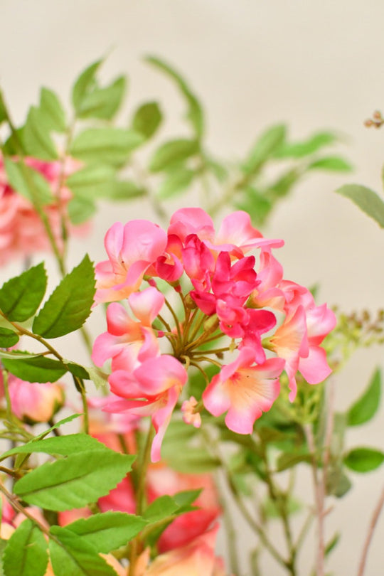 Elegantes Kunstblumenarrangement in warmen Rosa- und Koralltönen mit natürlicher, asymmetrischer Form und dekorativer Vase. Blumen Bahlmann München